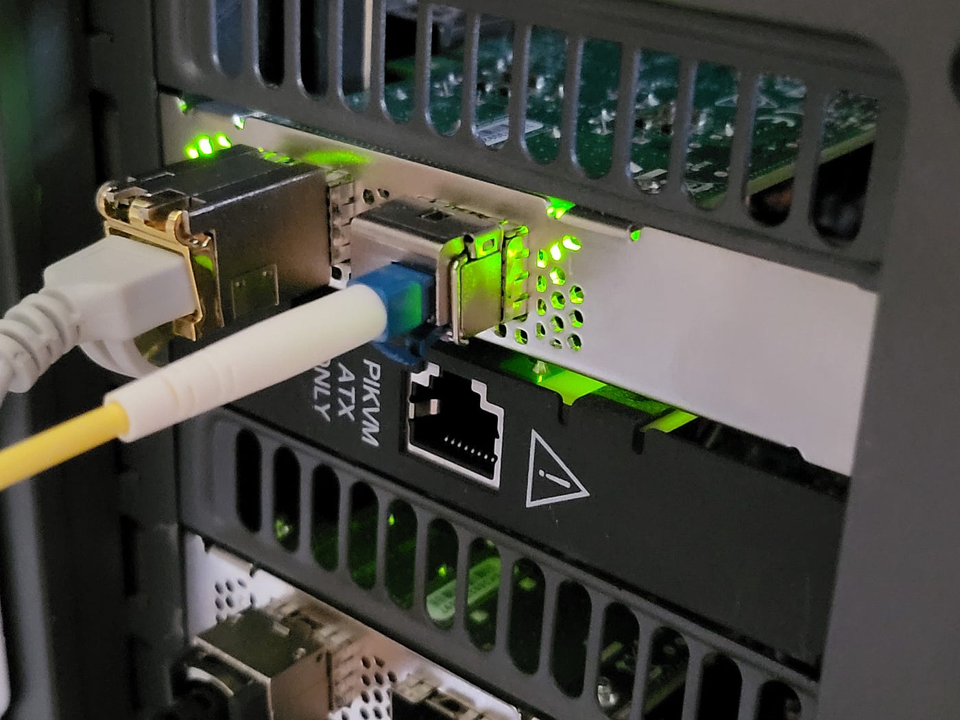 Backpanel of a router, close up, it shows SFP+ RJ45 with cat6e attached to it and SFP28 with fiber. Below — PiKVM4 addon panel but without any cables attached to it, and below, barely visible, another network card with DAC cable attached to it (second SFP28 port is free).