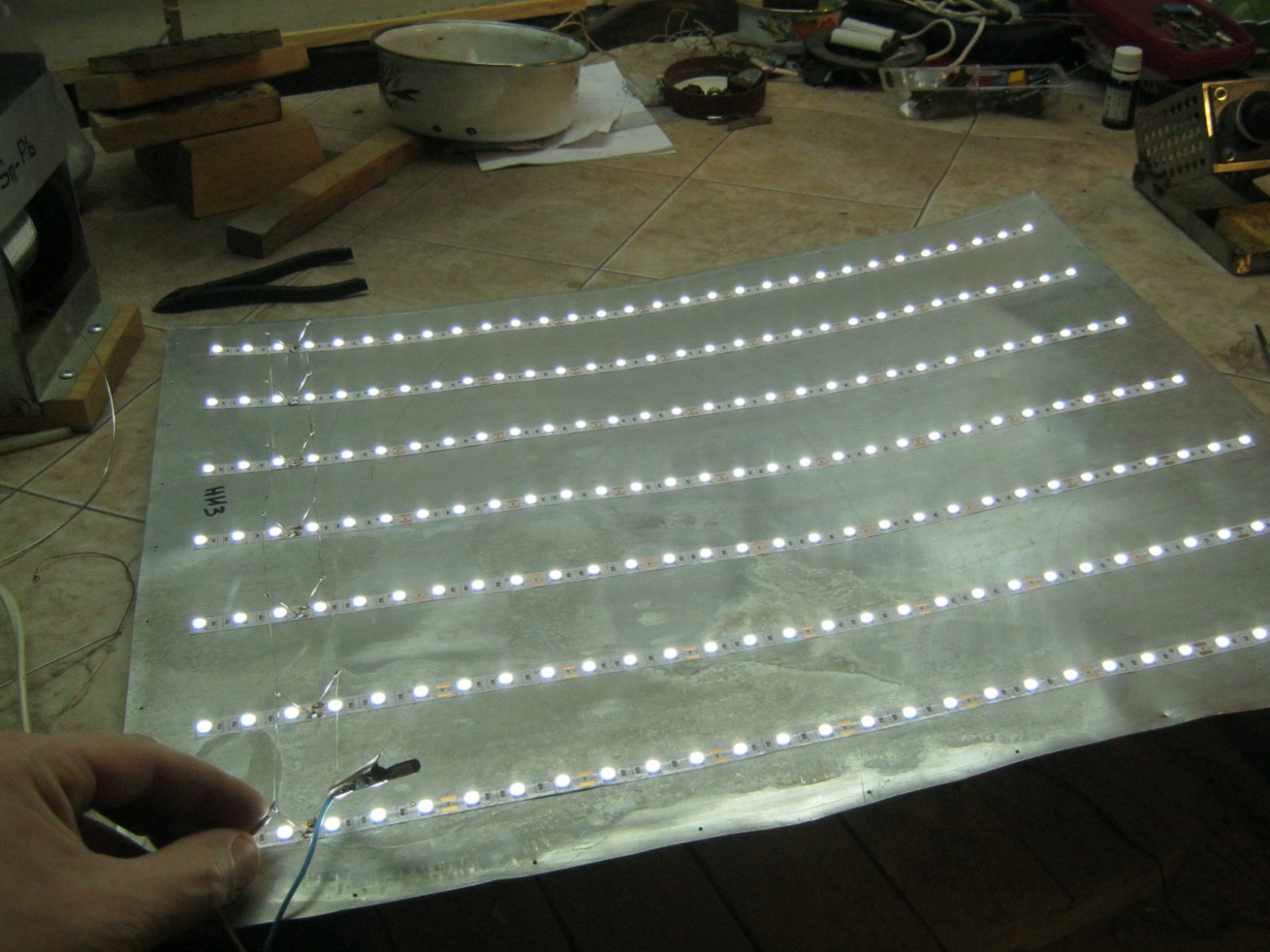 Фото 6.1. Лампа подсветки одного из светильников в сборе: из оцинкованной стали или алюминия ~0,5 мм толщиной, вырезана задняя стенка, размечена, обезжирена, наклеены нарезанные куски ленты с интервалом не более 50 мм. Отрезанные концы лент могут замыкать на металлическое основание, их следует подогнуть вверх и заизолировать лаком. Куски лент соединены параллельно, пайкой, нетолстым медным лужёным проводом — к каждой контактной площадке припаяны стойки, на их верху пинцетом сделаны крючочки, в которых зажаты и припаяны продольные шины. На фото — испытание лампы