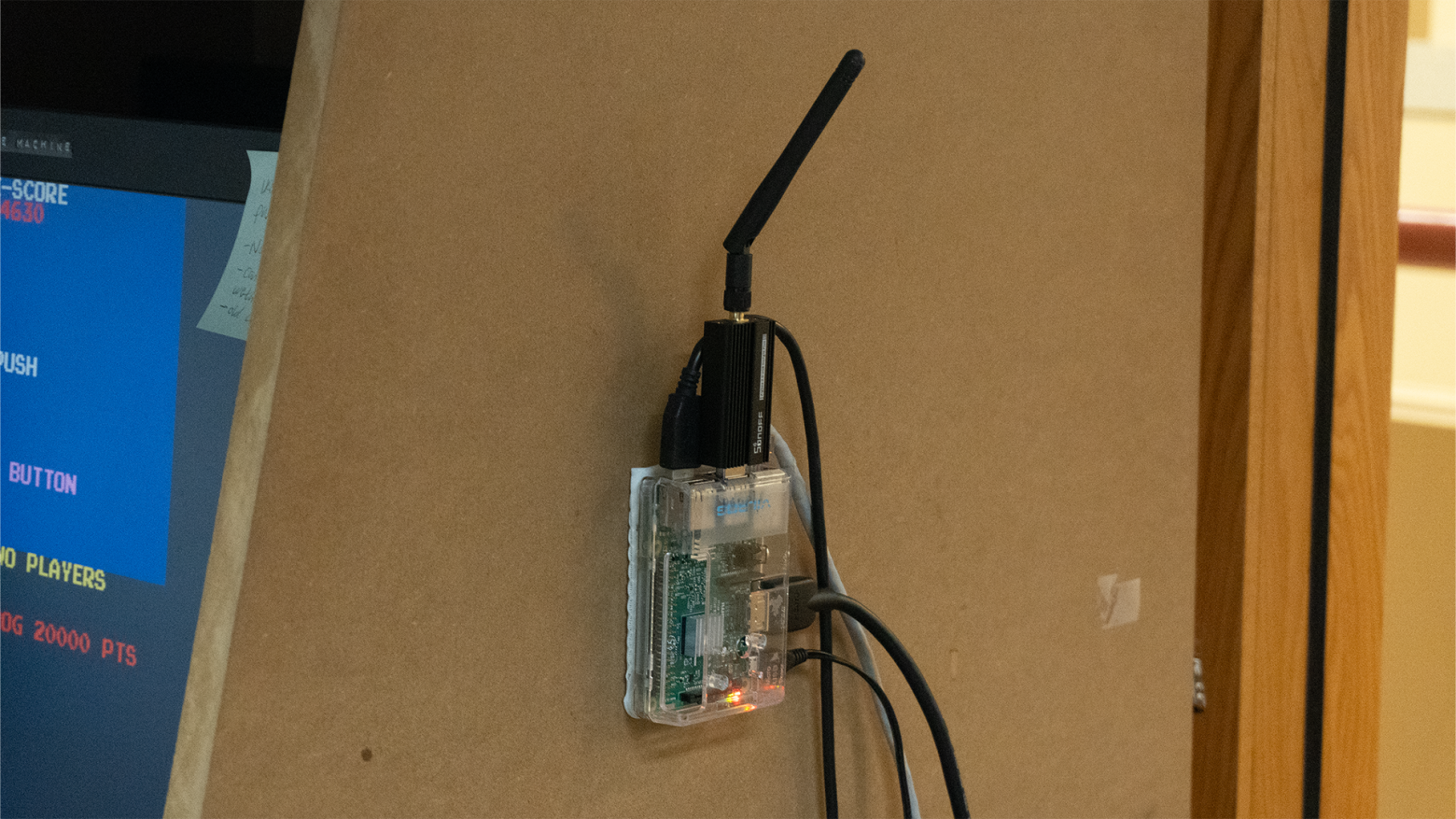 An image of a Raspberry Pi suspended from a wall with various cables plugged into it. There's a USB stick with an antenna sticking out.