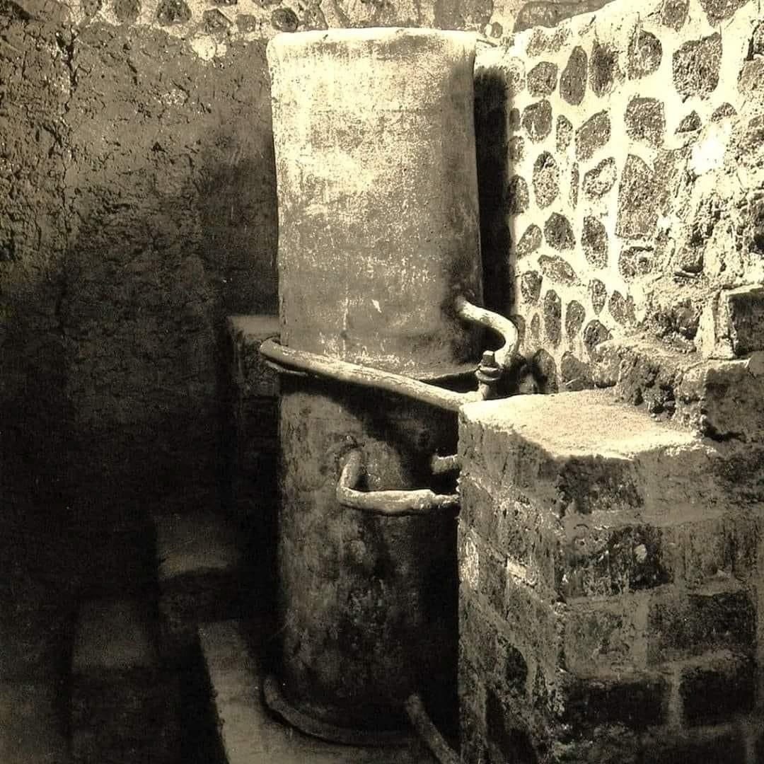 "Необыкновенная находка, найденная при раскопках виллы Пизанелла в Боскореале, недалеко от Помпей. К сожалению, это поселение не было объектом систематических раскопок, а только частичных исследований.Эти быстрые раскопки, проведенные без какой-либо научной систематики, имели целью извлечение драгоценных находок, которые сегодня разбросаны в различных музеях по всему миру.Первые свидетельства о вилле делла Пизанелла датируются ноябрем 1868 года, когда Модестино Пульцелла, прослеживая фундамент стены, обнаружил некие конструкции. Продолжая раскопки, была найдена мозаика. Затем раскопки пришлось прекратить, поскольку владелец соседней земли Винченцо Де Приско указал на опасность ущерба для его земли. Только в сентябре 1894 года раскопки были возобновлены, которые продолжались до июня 1895 года. На этот раз именно по инициативе Де Приско, который, очевидно, больше не боялся «ущерба».Так всплыло на свет знаменитое «сокровище Боскореале», сенсационно проданное за границу из-за пробела в законодательстве о защите культурного наследия. Был еще один перерыв, который длился около года, прежде чем раскопки возобновились в мае 1896 года.Так на свет появились римские бани и котел, найденные нетронутыми, со всеми их трубами. Затем Де Приско провел реставрацию части виллы, превратив ее в своего рода музей.И именно во время существования «Музея Де Приско» братья Алинари получили возможность сделать эту необычную фотографию"