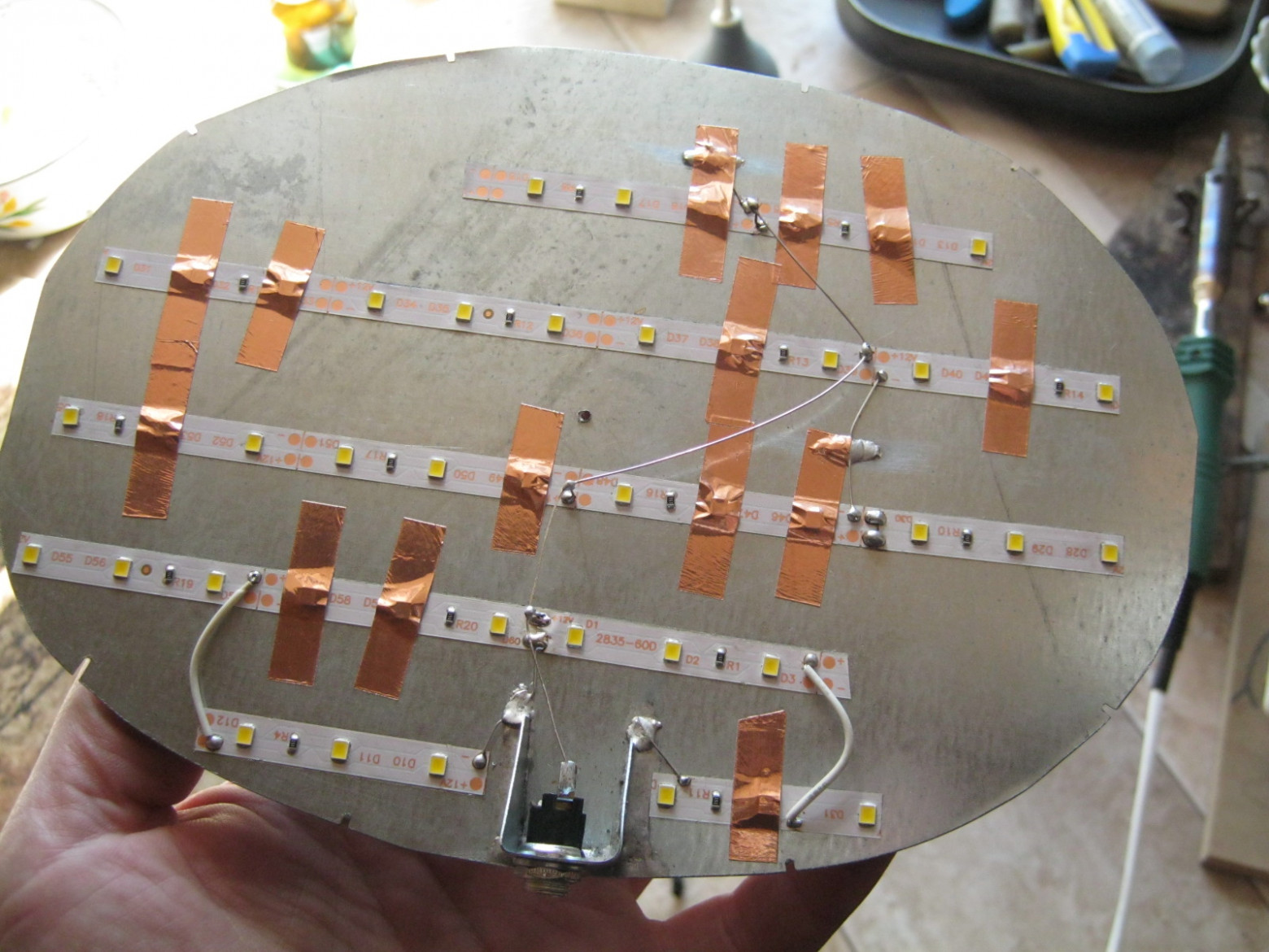 Фото 2.5. Кусочки белой, с нейтральным светом, светодиодной ленты, наклеиваем на обезжиренную пластину с внутренней стороны, согласно художественному замыслу работы. Стандартный низковольтный разъём от сетевого -12 В (-9В) адаптера удобно установить здесь же, на впаянном кронштейне. Соединяем отрезки ленты пайкой, тонким жёстким проводом, почаще примеряя лампу к стеклянной части. В качестве «—» питания удобно использовать «массу». Увы, в упрощённой открытой конструкции установка рассеивателя света затруднена, а точечный свет некоторых светодиодов заметен особенно сильно. Их можно закрыть непрозрачным, например, кусочком медной фольги с липким слоем — такой же, какую мы использовали при сборке витража Тиффани. Если фольгу не слишком сильно и тщательно обжимать вокруг светодиода, его выводов она не касается