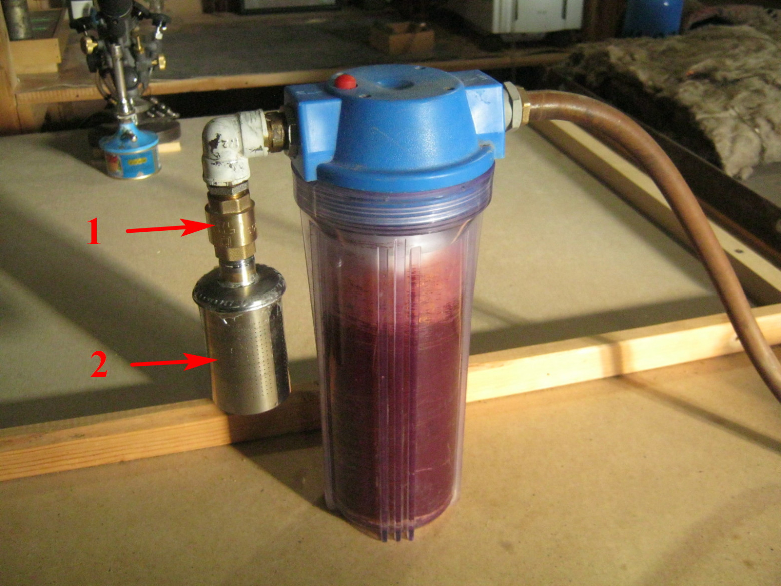 Фото 4.3. Внешний вид моего варианта осушителя. Б/у корпус спасён от свалки, мощный внутренний слой налёта ржавчины отчистил кухонной проволочной мочалочкой с горячей водой и содой. В верхней части, куда залезала моя лапа, это удалось получше, в нижней узкой части получилось не до прозрачности. В середину вставлен стандартный фильтр 10 микрон, со срезанным одним внешним слоем — чтобы освободить больше места для силикагеля и уменьшить сопротивление потоку воздуха. Верхние 2/3 фильтра обмотаны тонким упаковочным полиэтиленом, чтобы удлинить путь воздуха через силикагель. Последний засыпан между фильтрующим элементом и стенками колбы. Выход через переходник и штуцер соединён с мягким вакуумным шлангом, второй конец которого одет на входной патрубок концентратора. На входе осушителя обратный клапан 1 с удалённой пружиной. Тяжёлый металлический затвор при выключении концентратора падает в седло и отсекает силикагель от помещения. Противопылевой фильтр 2 спаял из нержавеющей сеточки от чайника и водопроводного хлама и набил синтепоном