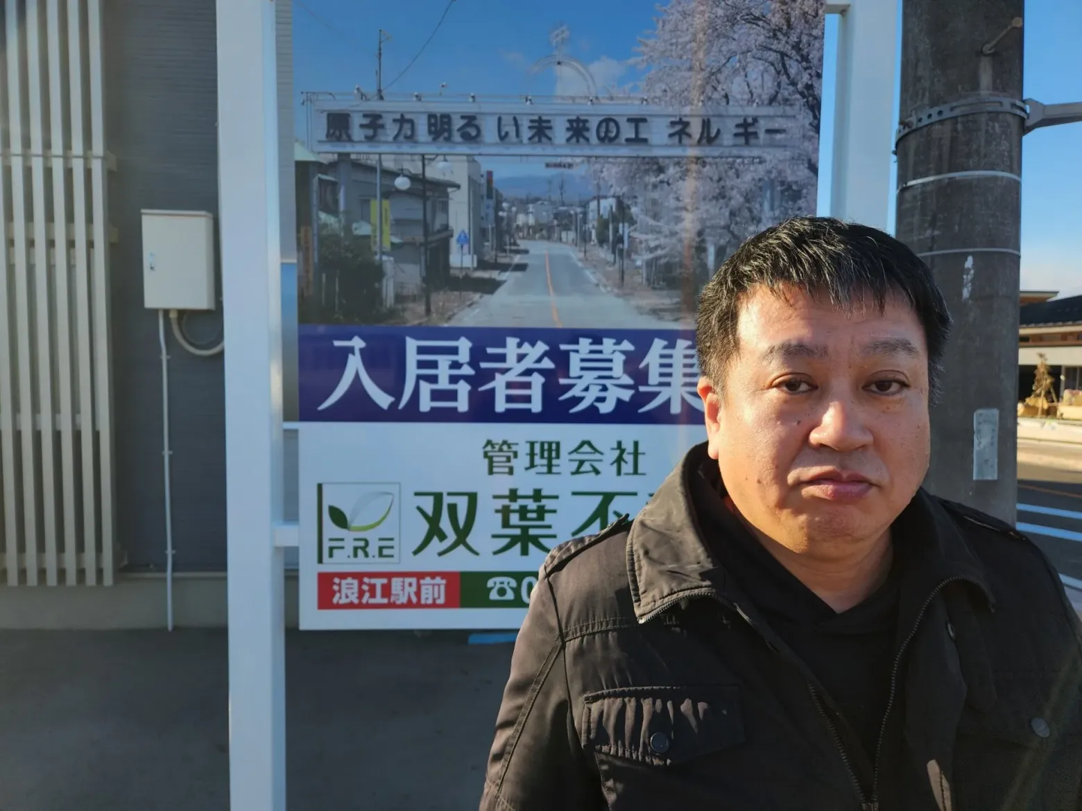 Yuji Onuma in front of his sign. The photo behind him shows the main street of Futaba, with the arch that Onuma designed as a schoolboy, which reads “Atomic power: Energy for a bright future.”  <span class="feature_caption_credit">Thomas A. Bass</span>