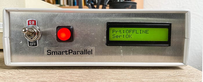 Photograph of the front panel of an electronic device with a large switch, a label that reads 'SmartParallel', an illuminated red button and an LCD display that shows the output: Prt: OFFLINE Ser: OK.